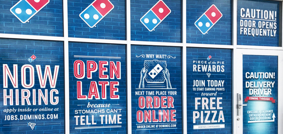 Storefront of Domino's featuring various promotional signs and hiring announcements.