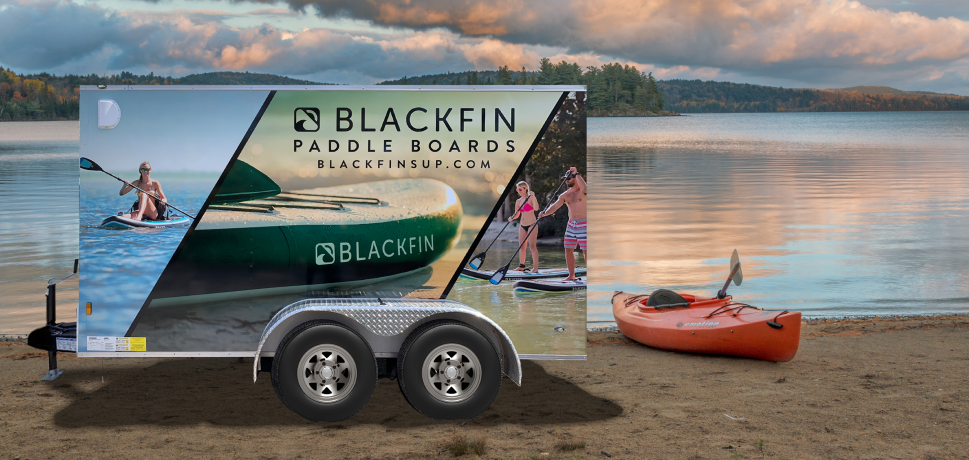 Trailer with Blackfin Paddle Boards branding parked near a lake with a kayak nearby.